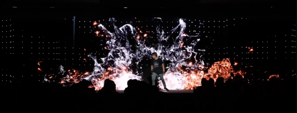 A person stands on a stage with a dynamic digital background displaying vivid abstract patterns of white and orange. Silhouetted audience members are visible in the foreground, enhancing the dramatic atmosphere.